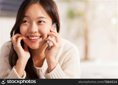 Asian smiling woman is phoning while lying on the sofa at home
