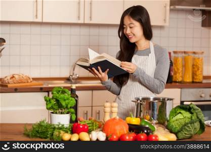 Asian smiling woman looking a cookbook while standing in her kitchen