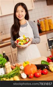 Asian smiling woman with a colorful salad in her hands is standing in the kitchen
