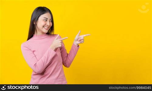 Asian smiling young woman finger Pointing to advertise, banner or showing on isolated yellow color background, wearing winter cozy sweater indoors studio, happiness and fun, copy space concept