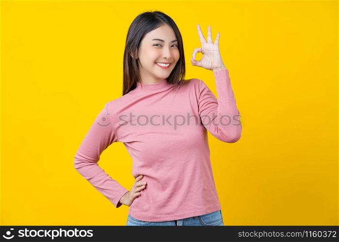 Asian smiling young woman gesturing ok sign for approval or agreement on isolated yellow color background, wearing winter cozy sweater indoors studio,happiness and optimistic decision copy space