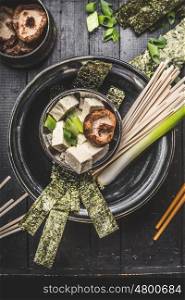 Asian soup preparation in bowl with udon noodls, tofu, Shiitake Mushrooms , nori and green onion on dark rustic background, top view