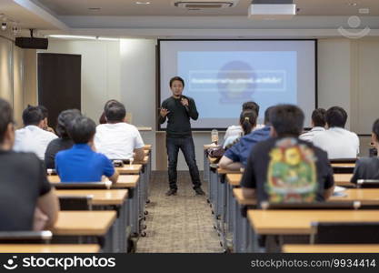 Asian Speaker with casual suit on the stage in front of the room with low light over the presentation screen in the business or education seminar, business and education concept