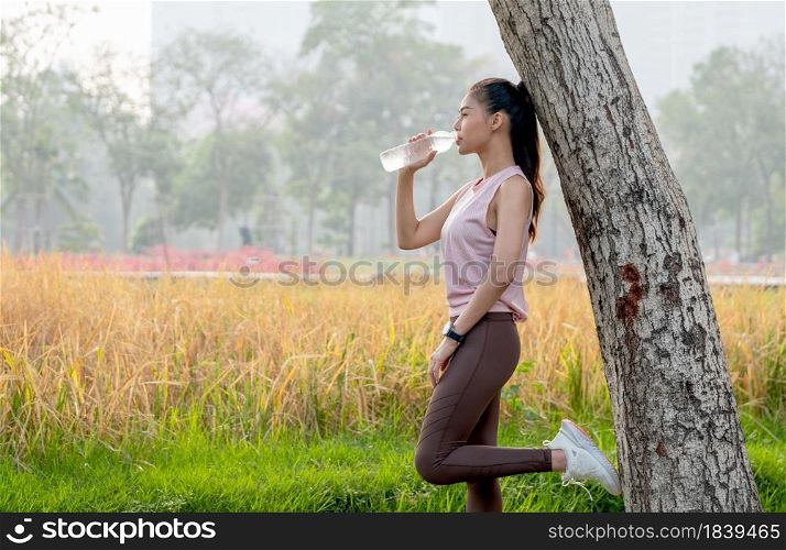 Asian sport woman lean tree and drink water from bottle with grass field as background during exercise in park or garden with morning light.