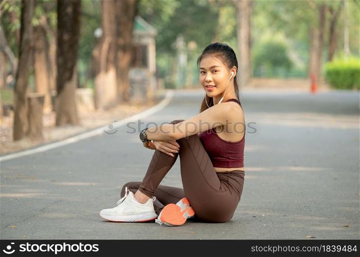 Asian sport woman sit and hug the knee also look at camera with smiling during exercise in park or garden in the morning.