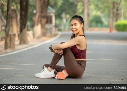 Asian sport woman sit and hug the knee also look at camera with smiling during exercise in park or garden in the morning.