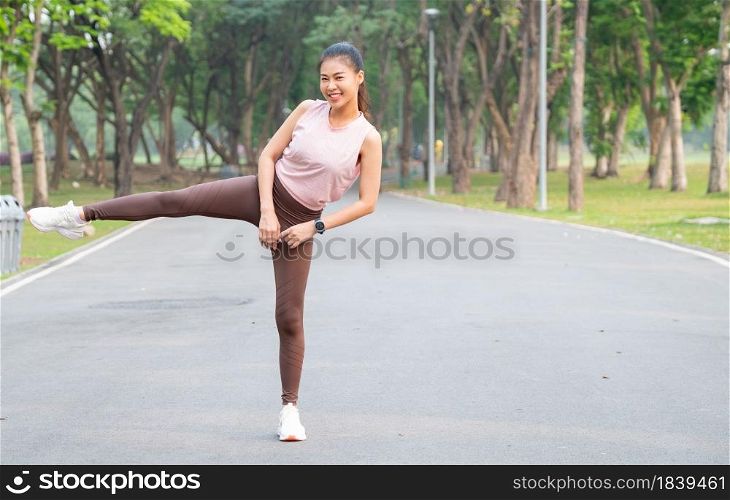Asian sport woman stand with action of stretch right leg to side also look at camera and smile on road of park or garden with morning light.