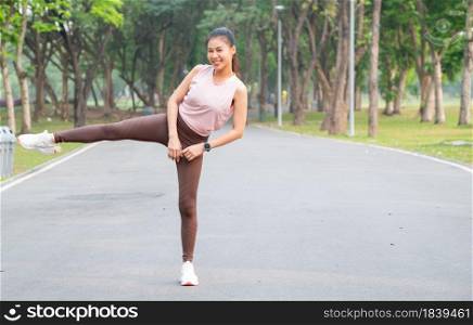 Asian sport woman stand with action of stretch right leg to side also look at camera and smile on road of park or garden with morning light.