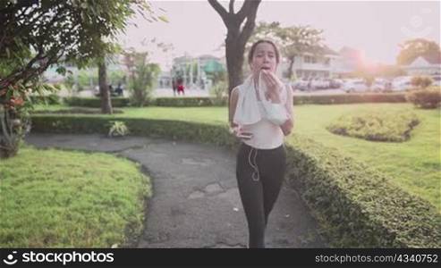 asian sporty woman using smartphone while walking down on the walking path at the recreation outdoor park during sunset hour, relaxing after workout, wiping sweat from face, new normal modern life