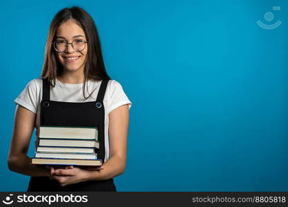 Asian student girl on blue background in the studio holds stack of university books from library. Woman smiles, she is happy to graduate. Copy space. Asian student girl on blue background in the studio holds stack of university books from library. Woman smiles, she is happy to graduate.