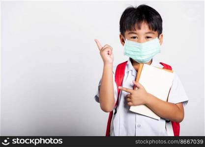 Asian student kid boy wear student thai uniform and protect face mask ready to go to school pointing finger to space in studio shot isolated on white background, preschool, new normal back to school