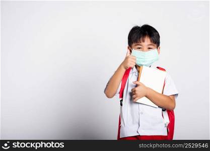 Asian student kid boy wearing student thai uniform and medical protect face mask and show thumb up finger for good in studio shot isolated on white background, new normal back to school