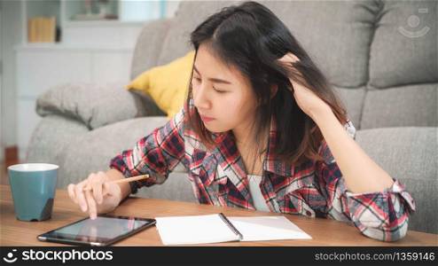 Asian student woman do homework at home, female drinking coffee and using tablet for searching on sofa in living room at home. Lifestyle women relax at home concept.