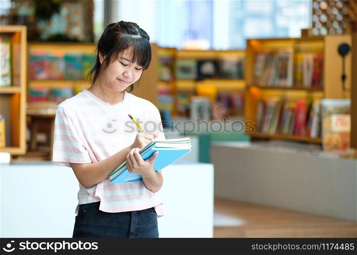 Asian teen girl student holding and writing her notebook standing over blur library with people background, education concept