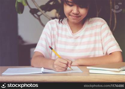Asian teenager girl student, with smiling face, writing notebook paper on table in her classroom, education concept