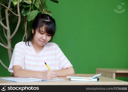 Asian teenager girl student, with smiling face, writing notebook paper on table in her classroom, education concept