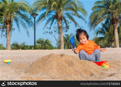 Asian Thai happy cute little cheerful daughter girl funny digging playing toy with sand at an outdoor tropical beach in summer day with copy space