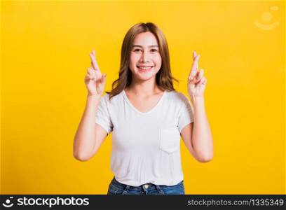 Asian Thai happy portrait beautiful cute young woman smile have superstition her holding fingers crossed for good luck and looking to camera, studio shot isolated on yellow background with copy space