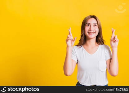 Asian Thai happy portrait beautiful cute young woman smile have superstition her holding fingers crossed for good luck and looking to camera, studio shot isolated on yellow background with copy space