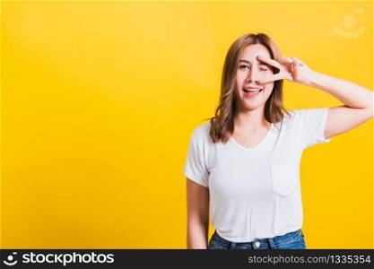 Asian Thai happy portrait beautiful cute young woman smile standing wear t-shirt showing finger making v-sign symbol near eye looking to camera, isolated studio shot yellow background with copy space