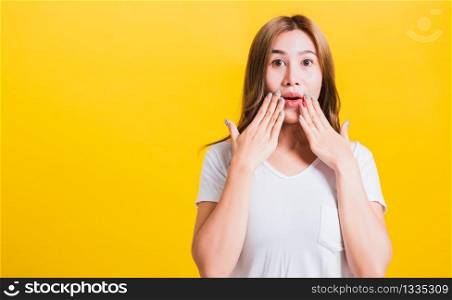 Asian Thai happy portrait beautiful cute young woman standing amazed, shocked afraid wide open mouth gesturing hand palms looking to camera, studio shot isolated on yellow background with copy space