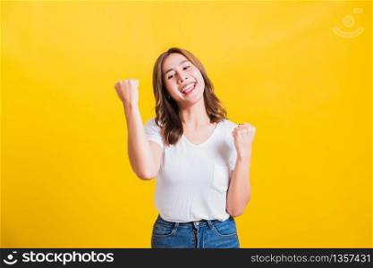 Asian Thai happy portrait beautiful cute young woman standing wear t-shirt makes raised fists up celebrating her winning success looking to camera isolated, studio shot on yellow background with copy space