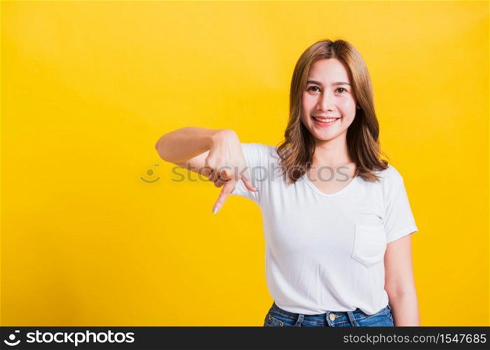 Asian Thai happy portrait beautiful cute young woman standing wear t-shirt makes gesture two fingers point below down and looking to camera, studio shot isolated on yellow background with copy space