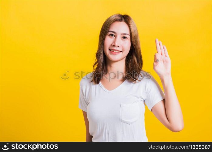 Asian Thai happy portrait beautiful cute young woman standing wear t-shirt showing gesturing ok sign with fingers looking to camera, isolated studio shot on yellow background with copy space