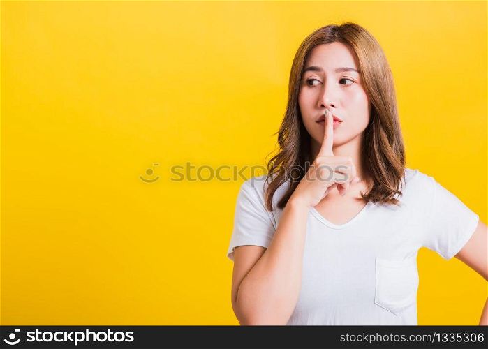 Asian Thai happy portrait beautiful cute young woman standing wear white t-shirt making finger on lips silent quiet gesture looking to side, studio shot isolated on yellow background with copy space