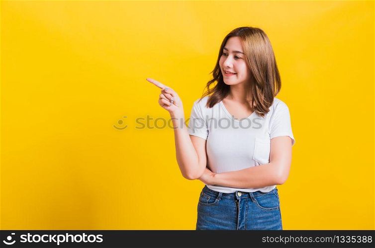 Asian Thai happy portrait beautiful cute young woman standing wear white t-shirt pointing finger away side looking to away side, studio shot isolated on yellow background with copy space