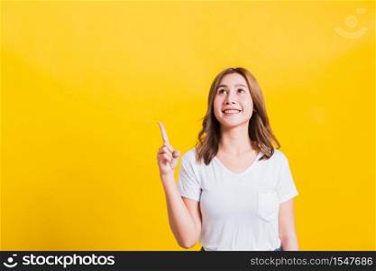 Asian Thai happy portrait beautiful cute young woman standing wear white t-shirt makes gesture two fingers point upwards above looking above, studio shot isolated on yellow background with copy space