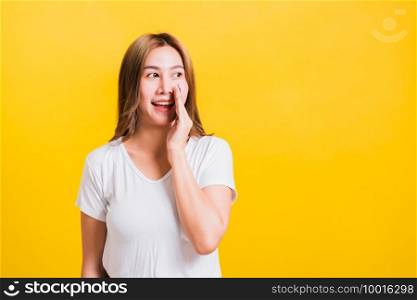 Asian Thai happy portrait beautiful young woman standing wear t-shirt hold a hand on mouth talking whispering secret rumor looking to camera isolated, studio shot on yellow background with copy space