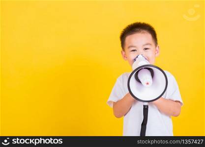 Asian Thai happy portrait cute little cheerful child boy holding and shouting or screaming through the megaphone her looking to camera, studio shot isolated on yellow background with copy space