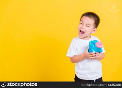 Asian Thai happy portrait cute little cheerful child boy smile holding piggy bank and looking camera, studio shot isolated on yellow background with copy space