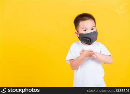 Asian Thai happy portrait cute little cheerful child boy wearing mask protective for covid-19 or pm2.5 dust he clean hand with sanitizing gel, studio shot isolated on yellow background with copy space