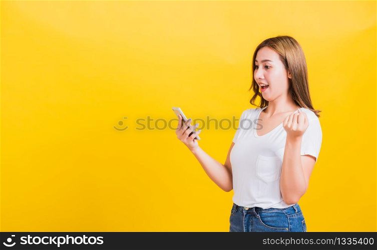 Asian Thai portrait happy beautiful cute young woman smiling stand wear t-shirt lift hand celebrating winning with smart mobile phone looking the phone isolated, on yellow background with copy space