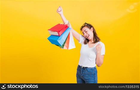 Asian Thai portrait happy beautiful cute young woman smiling stand with sunglasses excited holding shopping bags multi color looking camera, studio shot isolated yellow background with copy space