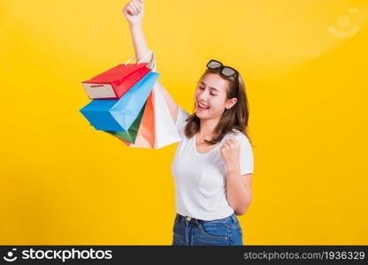 Asian Thai portrait happy beautiful cute young woman smiling stand with sunglasses excited holding shopping bags multi color looking camera, studio shot isolated yellow background with copy space