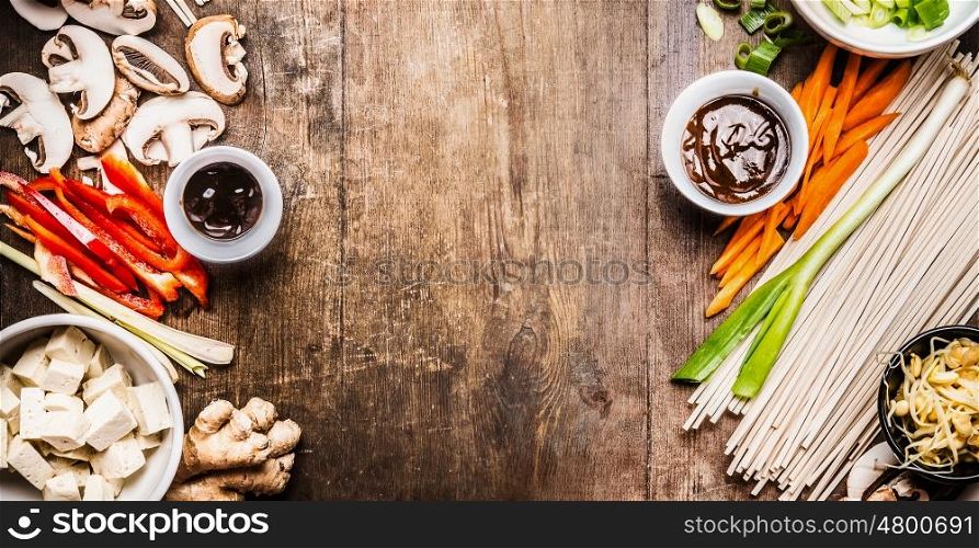 Asian vegetarian cooking ingredients for stir fry with tofu, noodles , vegetables and sauces on wooden rustic background, top view, banner, frame