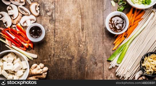 Asian vegetarian cooking ingredients for stir fry with tofu, noodles , vegetables and sauces on wooden rustic background, top view, banner, frame