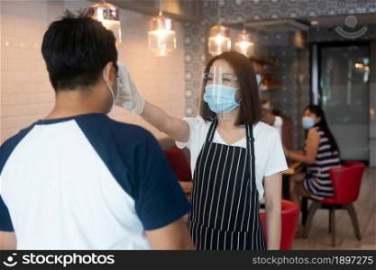 Asian waitress woman wearing face masks and holding an infrared forehead thermometer to check body temperature for virus symptoms of customers before entering the restaurant ( coffee shop ).