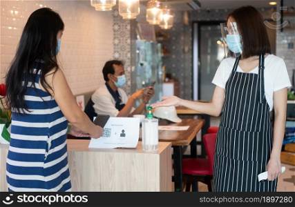 Asian waitress woman wearing face masks and holding an infrared thermometer and recommend customers to use hand alcohol sanitizer before entering the restaurant ( coffee shop ).