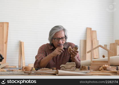 Asian white haired senior carpenter man holding magnifying glass to check completion of small Christmas tree model, wooden house, cup and toy car he made at home workshop. White background