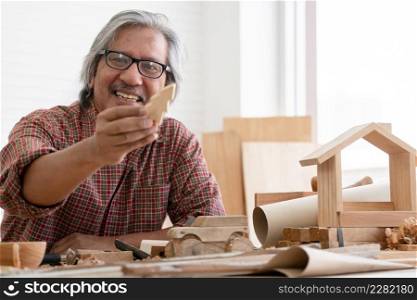 Asian white haired senior carpenter man smiling and showing small wooden Christmas tree model he made with pride at workplace at home. White background