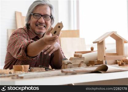 Asian white haired senior carpenter man smiling and showing small wooden house model he made with pride at workplace at home. White background