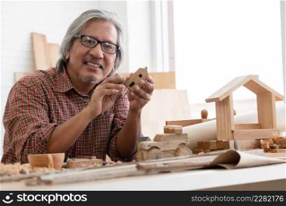 Asian white haired senior carpenter man smiling and using sandpaper to smooth the wooden model. Christmas tree model, wooden house, cup and toy car he made at home workshop. White background
