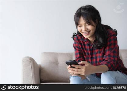 Asian woman are using a smartphone while on sitting sofa in the living room at home. Woman wearing a red plaid shirt look social media through mobile phone