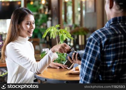 Asian woman customer using credit card swipe on credit card reader EDC machine to pay a waiter for coffee purchase at table in cafe.