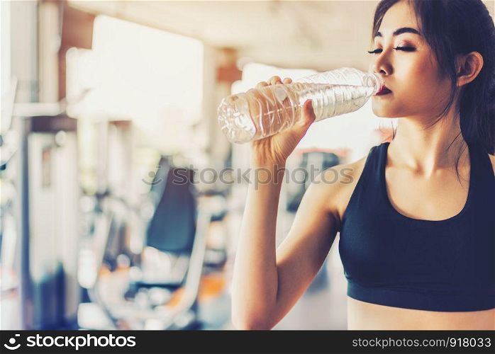 Asian woman drinking pure drinking water for freshness after workout or exercise training in fitness gym with fitness equipment background. Relax and Rest concept. Strength and Fitness training theme.