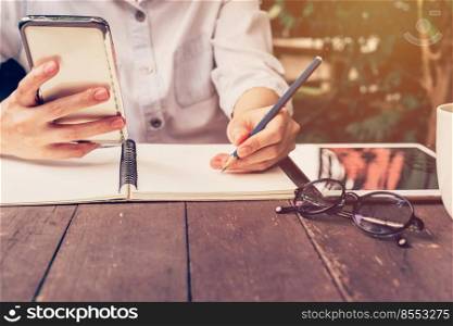 asian woman hand holding phone and pencil writing notebook in coffee shop with vintage toned